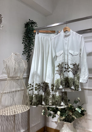 A white two-piece linen set, a long-sleeve button-up shirt and matching skirt, both decorated with a green palm tree print. 